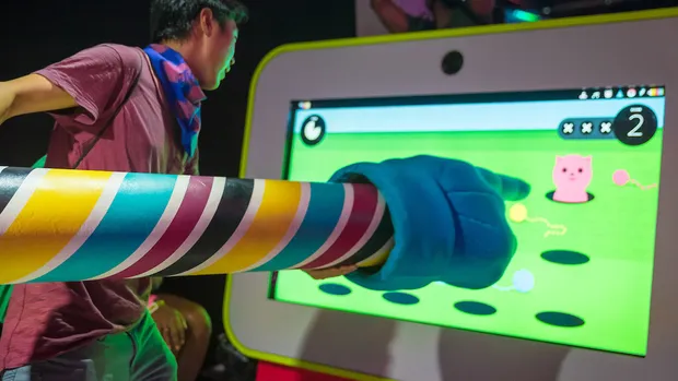 A giant foam hand poking a big screen with a game on it.